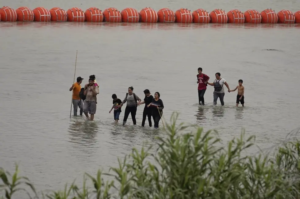 Migrantes que cruzaron el río Grande (Bravo) desde México caminan frente a enormes boyas colocadas como una barrera fronteriza flotante en el río, el miércoles 12 de julio de 2023, en Eagles Pass, Texas. (AP Foto/Eric Gay)