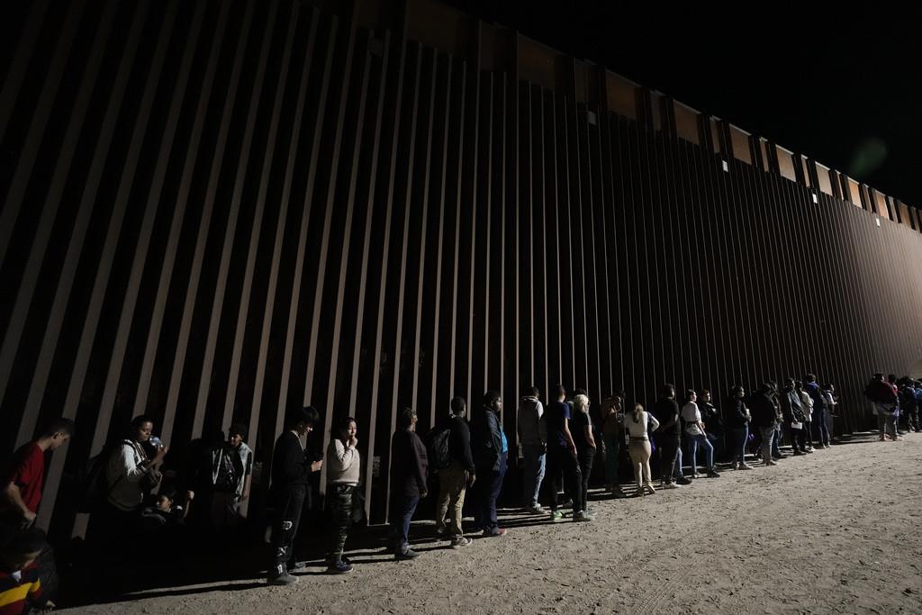 Varias personas se forman junto a un muro fronterizo mientras esperan solicitar asilo tras cruzar la frontera desde México, el martes 11 de julio de 2023, cerca de Yuma, Arizona. (AP Foto/Gregory Bull, Archivo)